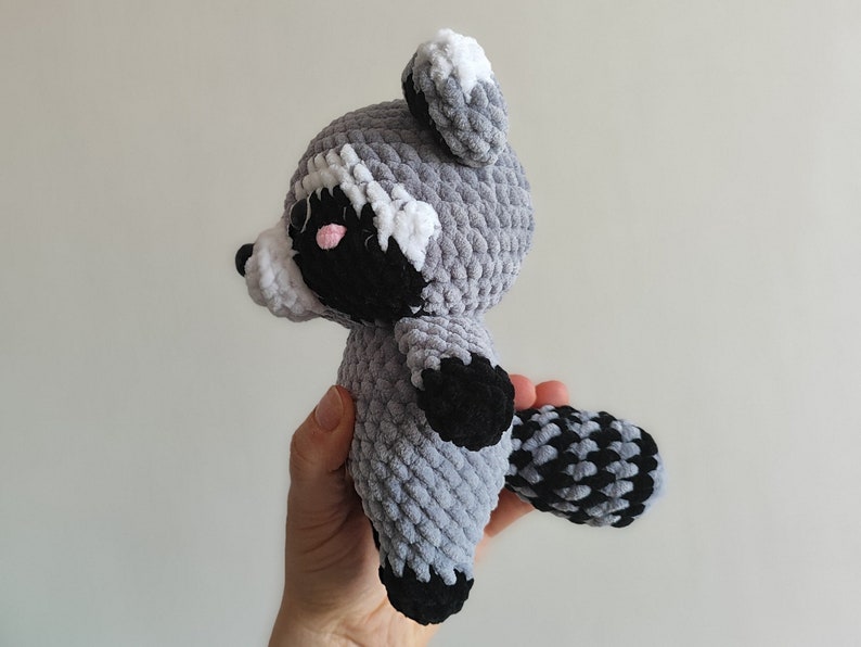 May include: A crocheted raccoon plush toy with gray and black fur, white ears, and pink cheeks. The toy is being held by a hand.