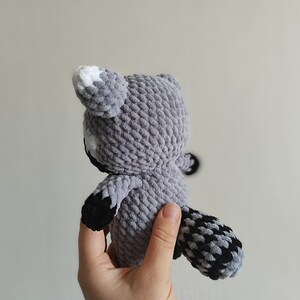 May include: A gray crocheted raccoon plush toy with black and white accents. The toy is being held by a hand.