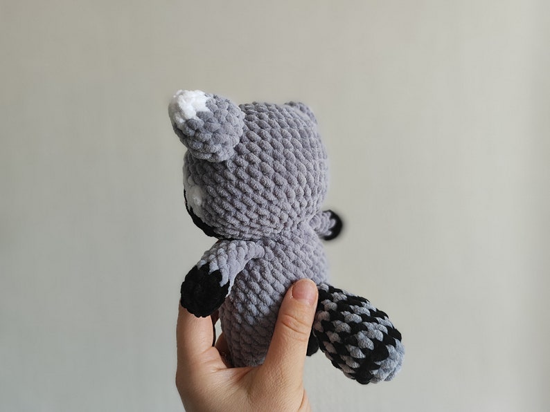 May include: A gray crocheted raccoon plush toy with black and white accents. The toy is being held by a hand.