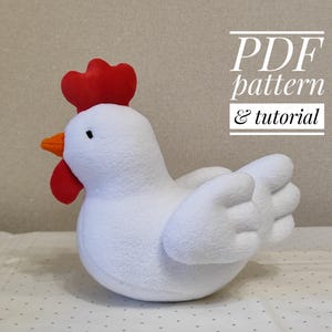 Peut inclure: Un jouet en peluche de poulet blanc avec une crête et un bec rouges. Le jouet est assis sur une surface blanche. Le texte "PDF pattern & tutorial" est visible en arrière-plan.