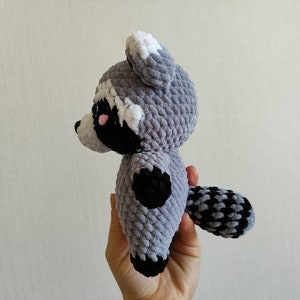 May include: A gray and black crocheted raccoon plush toy with white ears and a black nose. The toy is being held by a hand.