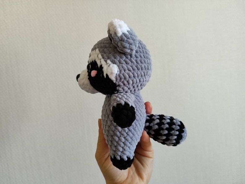 May include: A gray and black crocheted raccoon plush toy with white ears and a black nose. The toy is being held by a hand.