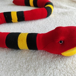 May include: A plush snake toy in red, black, and yellow. The snake's body is primarily red with black and yellow bands. The head is red with black eyes and a yellow mouth and tongue. The toy is lying on a white, textured surface.