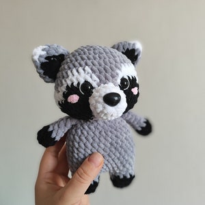 May include: A crocheted plush raccoon toy with gray and white fur, black eyes, a black nose, and pink cheeks.