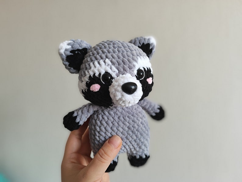 May include: A crocheted plush raccoon toy with gray and white fur, black eyes, a black nose, and pink cheeks.
