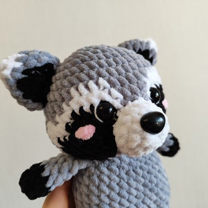 May include: A gray and white crocheted raccoon plush toy with black eyes and a black nose. The toy has pink cheeks and a white stripe on its forehead.