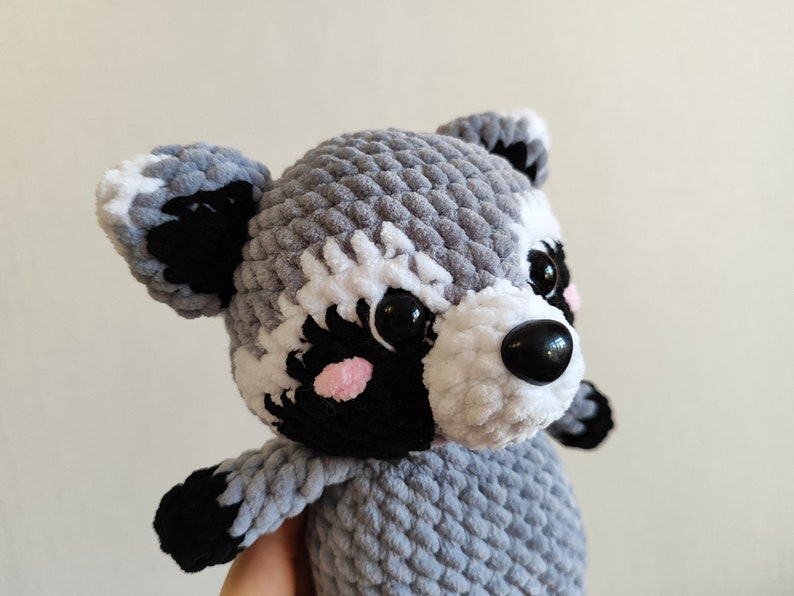 May include: A gray and white crocheted raccoon plush toy with black eyes and a black nose. The toy has pink cheeks and a white stripe on its forehead.