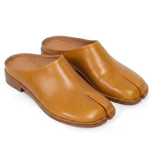 Op de afbeelding: Een paar bruinleren instappers met een split-teenontwerp. De schoenen hebben een lage hak en een gladde, gepolijste afwerking. De schoenen staan tegen een witte achtergrond.
