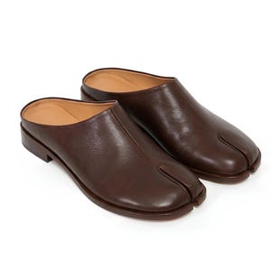 May include: A pair of brown leather split-toe mules. The shoes have a slip-on design with a low heel and a distinctive split between the big toe and the other toes. The interior is a lighter shade of brown.