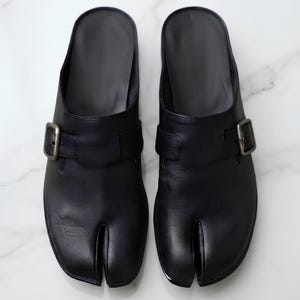 May include: A pair of black leather split-toe mules featuring a wide strap across the top with an adjustable antique brass-toned metal buckle. The footwear has a smooth finish, a rounded silhouette, and a distinct cleft design at the front toe area.