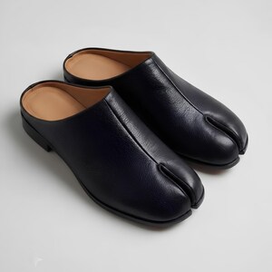 May include: A pair of black leather slip-on mules featuring a distinct split-toe design. The shoes have a textured grain finish on the upper, a smooth tan interior lining, and a low heel, showcasing a minimalist construction with a central seam.