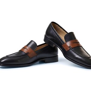 May include: A pair of black leather penny loafers featuring a contrasting tan leather strap across the vamp. The shoes have a low heel, a rounded toe, and a smooth finish. One shoe is positioned flat while the other rests at an angle.