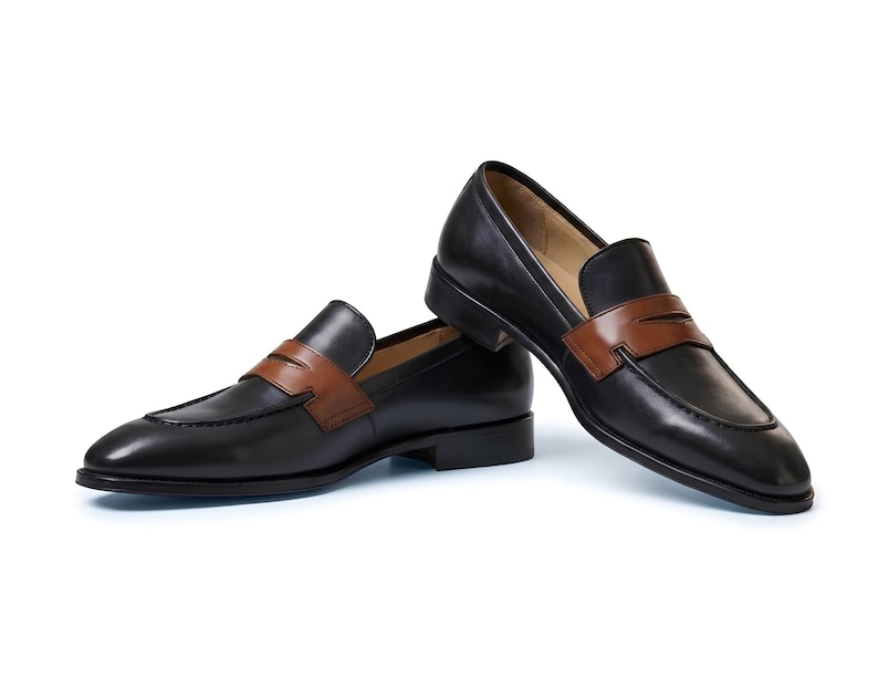 May include: A pair of black leather penny loafers featuring a contrasting tan leather strap across the vamp. The shoes have a low heel, a rounded toe, and a smooth finish. One shoe is positioned flat while the other rests at an angle.