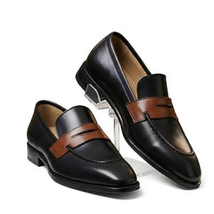 May include: A pair of black leather loafers featuring a contrasting brown leather saddle strap across the vamp. The shoes have a smooth finish, a rounded toe, and a low heel. One shoe rests on the ground while the other is elevated on a clear stand.
