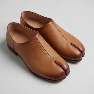 May include: A pair of tan leather shoes with a distinct split-toe design. The shoes feature a textured grain finish, a low-cut silhouette, and a smooth interior lining. The construction includes a central seam running from the split toe to the collar.