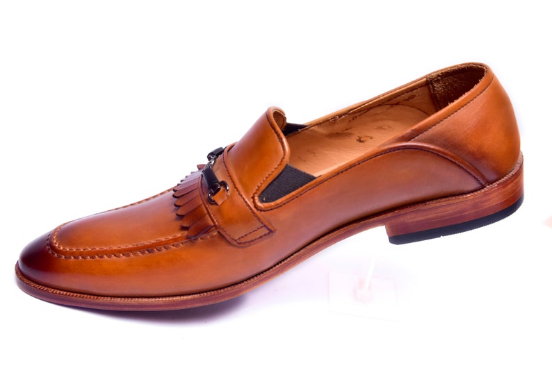 K&ouml;nnte beinhalten: Ein brauner Leder-Slipper mit Quaste und einer Metallschnalle. Der Schuh hat eine braune Ledersohle und einen schwarzen Absatz.