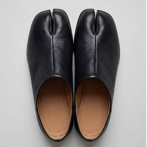 May include: A pair of black leather shoes featuring a distinct split-toe design. The shoes have a textured grain finish on the exterior, a smooth tan interior lining, and a low-profile silhouette. The design separates the big toe from the rest.