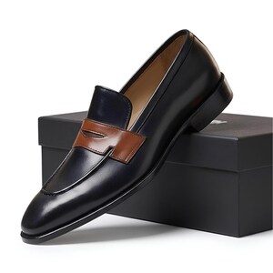 May include: A dark navy blue leather penny loafer with a smooth finish and a contrasting tan leather saddle strap across the vamp. The shoe features a rounded toe, a low stacked heel, and a tan interior lining, resting on a black rectangular box.