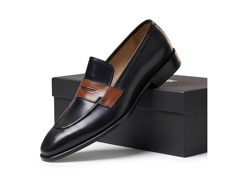 May include: A dark navy blue leather penny loafer with a smooth finish and a contrasting tan leather saddle strap across the vamp. The shoe features a rounded toe, a low stacked heel, and a tan interior lining, resting on a black rectangular box.