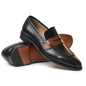 May include: A pair of black leather loafers featuring a contrasting brown leather saddle strap across the vamp. The shoes have a low heel, a pointed toe, and a tan interior lining. The sole of the shoe is partially brown with visible metal studs.