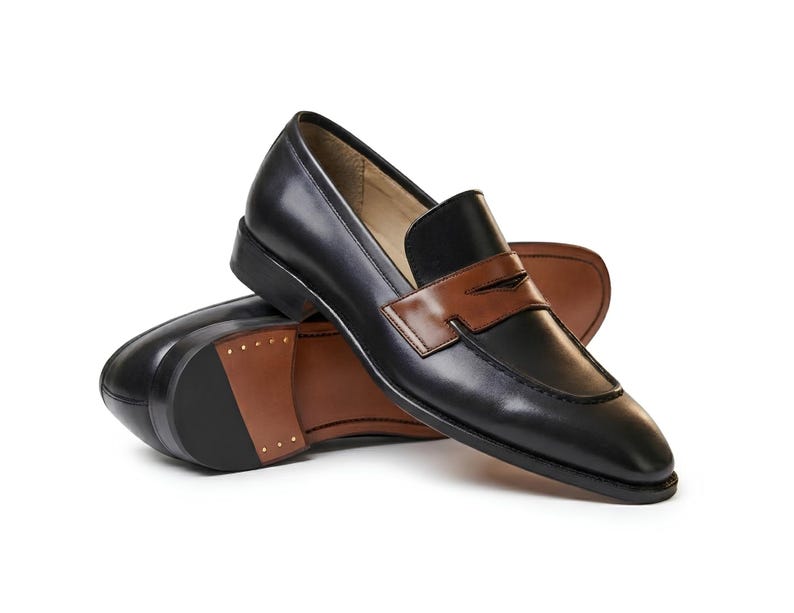 May include: A pair of black leather loafers featuring a contrasting brown leather saddle strap across the vamp. The shoes have a low heel, a pointed toe, and a tan interior lining. The sole of the shoe is partially brown with visible metal studs.