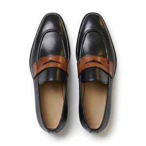 May include: A pair of black leather loafers featuring a smooth finish and a contrasting tan leather strap across the vamp. The shoes have a rounded toe design, a low profile, and a light-colored interior lining, presented from a top-down view.