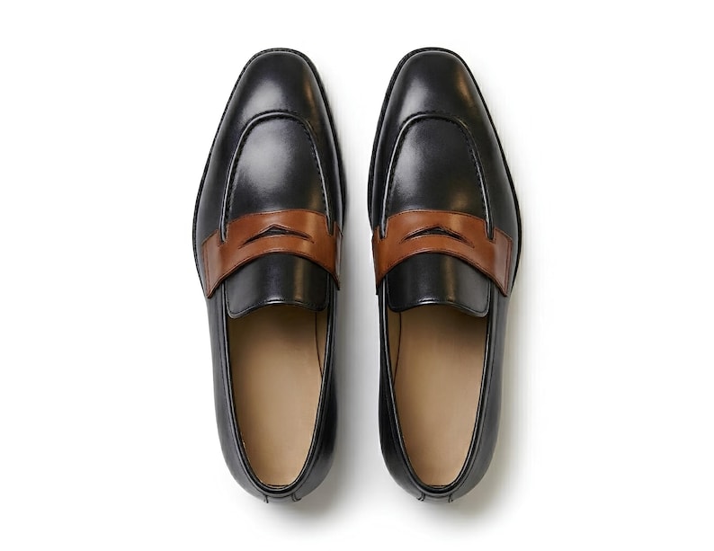 May include: A pair of black leather loafers featuring a smooth finish and a contrasting tan leather strap across the vamp. The shoes have a rounded toe design, a low profile, and a light-colored interior lining, presented from a top-down view.