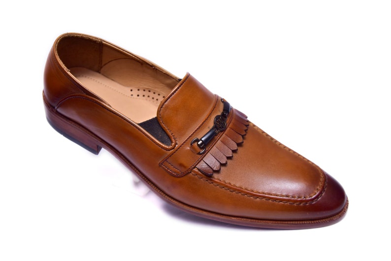 K&ouml;nnte beinhalten: Ein brauner Leder-Slipper mit Fransen-Detail und einer Metallschnalle. Der Slipper hat eine spitze Zehenpartie und eine Ledersohle.