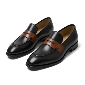May include: A pair of black leather loafers featuring a smooth finish and a contrasting brown leather strap across the vamp. These slip-on shoes have a rounded toe, a low heel, and a tan interior lining, showcasing a classic two-tone design.