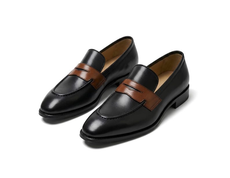 May include: A pair of black leather loafers featuring a smooth finish and a contrasting brown leather strap across the vamp. These slip-on shoes have a rounded toe, a low heel, and a tan interior lining, showcasing a classic two-tone design.