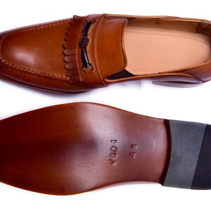 K&ouml;nnte beinhalten: Ein Paar braune Lederschuhe mit Quaste und einer Metallschnalle. Die Schuhe haben eine Ledersohle und sind Gr&ouml;&szlig;e 41.