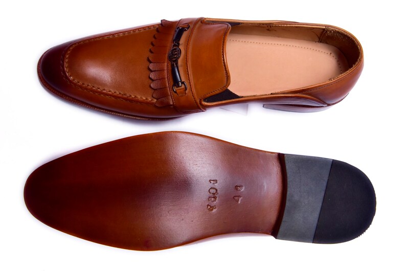 K&ouml;nnte beinhalten: Ein Paar braune Lederschuhe mit Quaste und einer Metallschnalle. Die Schuhe haben eine Ledersohle und sind Gr&ouml;&szlig;e 41.