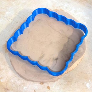 Könnte beinhalten: Ein blauer Ausstecher mit gewelltem Rand liegt auf einem Tonklumpen. Der Ton befindet sich auf einem Holzschneidebrett. Ein Nudelholz und Holzleisten sind im Hintergrund.