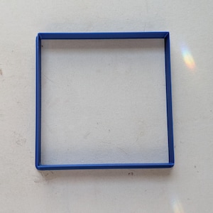 May include: A square, blue frame sits on a white, textured surface. The frame is a solid, vibrant blue, and the surface has subtle imperfections. Light refracts, creating a rainbow effect.
