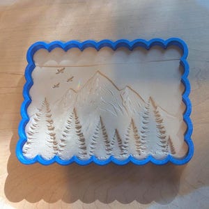 Puede incluir: Cortador de galletas con borde azul con un diseño de montaña y bosque. El cortador presenta un paisaje detallado con árboles, montañas y pájaros. El cortador es rectangular con un borde festoneado.