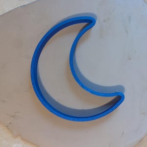 Może przedstawiać: Niebieska wykrawaczka w kształcie półksiężyca wciśnięta w jasnobrązowy kawałek gliny. Glina leży na jasnej drewnianej powierzchni. Wykrawaczka jest wykonana z tworzywa sztucznego.