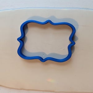 Może przedstawiać: Niebieska plastikowa foremka do ciastek w kształcie ozdobnej ramki lub nawiasu. Foremka jest wciśnięta w płaską, jasną powierzchnię ciasta, tworząc wyraźny zarys prostokątnego kształtu z zakrzywionymi, ozdobnymi krawędziami ze wszystkich stron.