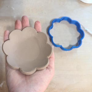 May include: A beige clay dish with a scalloped edge, held in a hand. A blue, scalloped-shaped cookie cutter is next to it. The dish is likely for small items or decoration. The items are on a wooden surface.