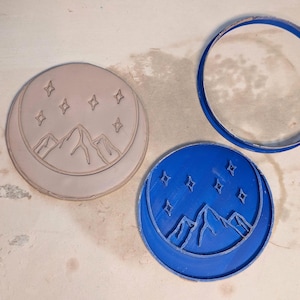 May include: Three cookie cutters: one light gray, one blue, and a blue ring. The gray cutter is a circle with a crescent moon and mountain design, with star shapes. The blue cutter has the same design. The blue ring is a circle.