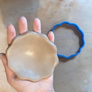 Puede incluir: Una forma de arcilla beige claro con bordes festoneados sostenida en una mano, junto a un cortador de galletas de plástico azul con un diseño festoneado a juego. La forma de arcilla parece recién formada y lista para hornear o crear.