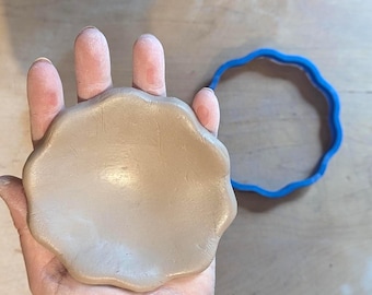 Può includere: Una forma di argilla beige chiaro con bordi smerlati tenuta in una mano, accanto a un tagliabiscotti di plastica blu con un design smerlato abbinato. La forma di argilla sembra appena modellata e pronta per la cottura o la creazione.