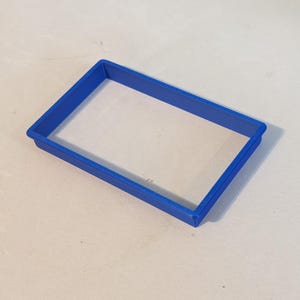Puede incluir: Un cortador de galletas rectangular de plástico azul cobalto. El cortador tiene un borde elevado y un centro hueco. El objeto está sobre una superficie de color claro.
