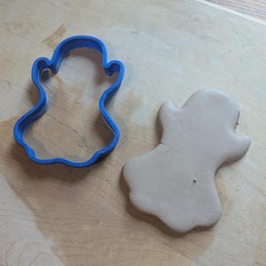 May include: A blue plastic ghost-shaped cookie cutter next to a light brown ghost-shaped piece of dough. The cutter is on a light wooden surface, and the dough is next to the cutter.