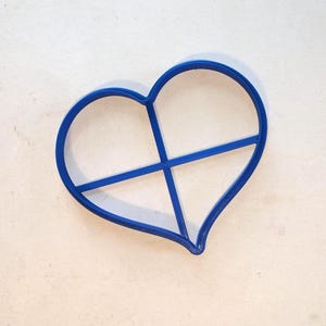 May include: A blue heart-shaped cookie cutter with a cross design. The plastic cutter sits on a light background. Suitable for baking or craft projects. The cutter's shape is clearly defined.