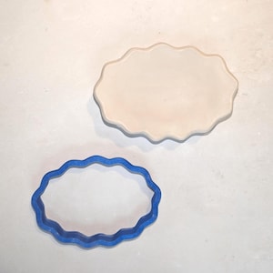 May include: A blue plastic oval-shaped cookie cutter with a scalloped edge and a matching light beige oval-shaped piece of clay or dough that has been cut into the same scalloped oval shape, both displayed together on a flat surface.