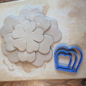 May include: A light brown clay flower design with heart-shaped petals, arranged on a wooden surface. A set of three blue heart-shaped cookie cutters are next to the clay flower. The clay flower is a craft project.
