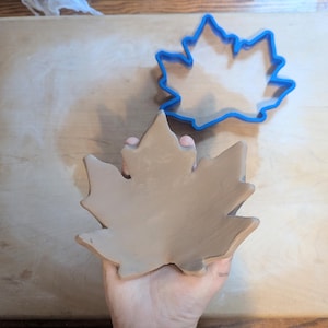 Puede incluir: Un plato en forma de hoja de arce de arcilla marrón claro, sostenido en una mano, junto a un cortador de galletas de hoja de arce de plástico azul. El plato está sobre una superficie de madera marrón claro.