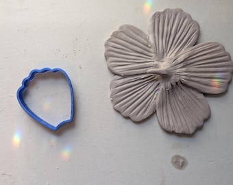 May include: A light gray, clay hibiscus flower next to a blue, petal-shaped cookie cutter. The flower has detailed petal lines and a textured surface. The cookie cutter is made of blue plastic.