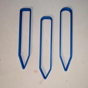 Puede incluir: Tres cortadores de galletas azules en forma de lápiz de diferentes tamaños. Cada cortador tiene una punta y una parte superior redondeada o cuadrada. Los cortadores están hechos de un material sólido y opaco.