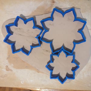 May include: Three blue flower-shaped cookie cutters of varying sizes are arranged on a light brown clay surface. The clay rests on a wooden surface, suggesting a crafting or baking project.
